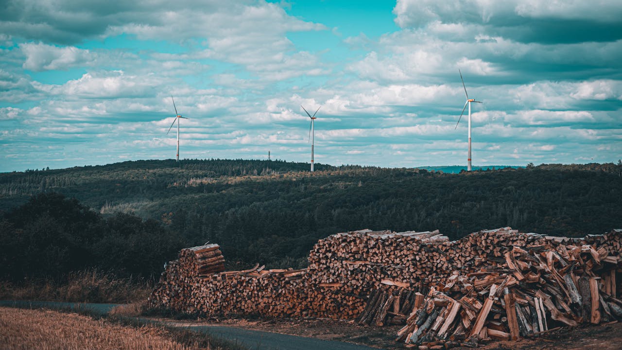 hero-img-02 Wind turbines on a hill with stacked logs in the foreground under a cloudy sky.