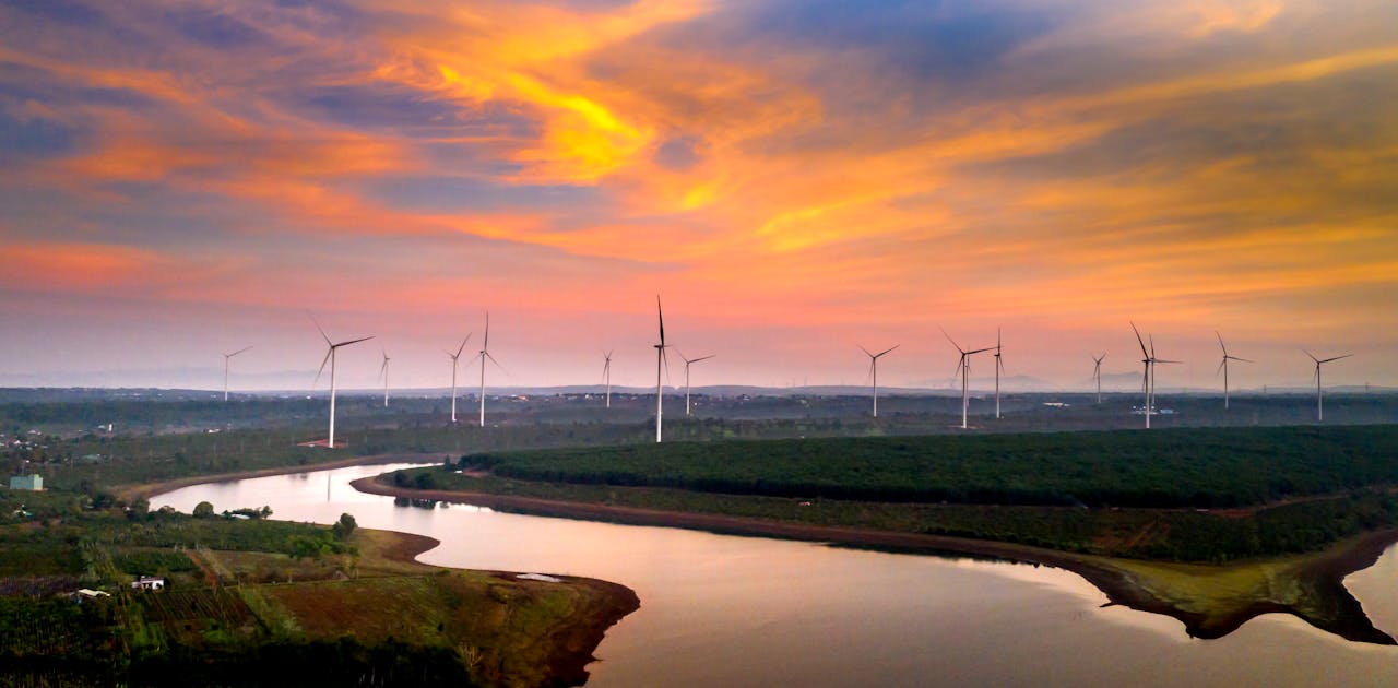 hero-img-01 Aerial view of wind turbines in a vibrant sunset sky over a river and fields.